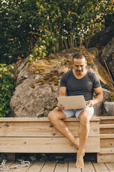 Reifer Mann in voller Länge, der einen Laptop benutzt, während er auf einer Terrasse vor Bäumen sitzt - MASF11101