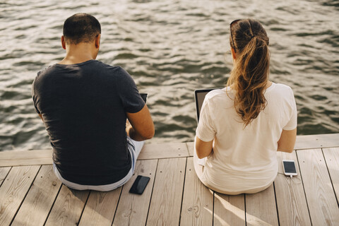 Rückansicht eines Paares, das einen Laptop benutzt, während es auf der Terrasse am See sitzt, lizenzfreies Stockfoto