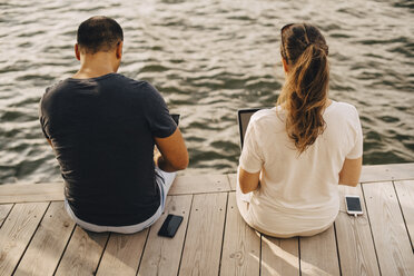 Rückansicht eines Paares, das einen Laptop benutzt, während es auf der Terrasse am See sitzt - MASF11106