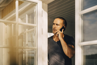 Thoughtful man talking on smart phone while standing at window - MASF11115