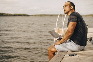 Thoughtful mature man sitting with laptop on jetty over lake - MASF11117
