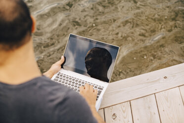 High angle view of smiling man reflecting on laptop at jetty - MASF11118