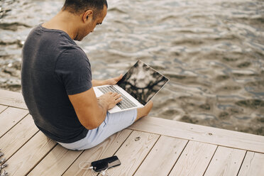 Älterer Mann benutzt Laptop, während er auf einem Steg über einem See sitzt - MASF11119