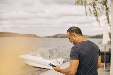 Älterer Mann, der ein Smartphone benutzt, mit einer Yacht, die im Hintergrund am See vertäut ist - MASF11121