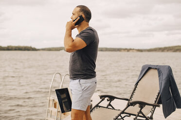 Mann mit Laptop, der mit seinem Smartphone spricht, während er auf einem Steg am See steht, gegen den Himmel - MASF11122