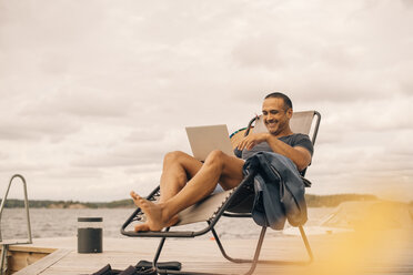 Smiling mature man using laptop while sitting on deck chair at jetty against cloudy sky - MASF11129