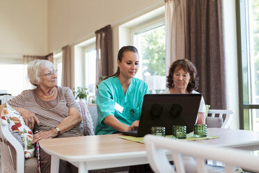 Junge Krankenschwester benutzt Laptop, während sie inmitten älterer Frauen im Pflegeheim sitzt - MASF11150