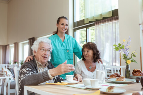 Eine lächelnde Krankenschwester und eine ältere Frau hören einem Mann beim Frühstück zu Hause zu, lizenzfreies Stockfoto