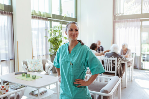 Lächelnde junge Altenpflegerin mit Hand auf der Hüfte im Pflegeheim stehend, lizenzfreies Stockfoto