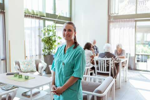 Porträt einer lächelnden Krankenschwester mit älteren Menschen im Hintergrund in einem Pflegeheim, lizenzfreies Stockfoto