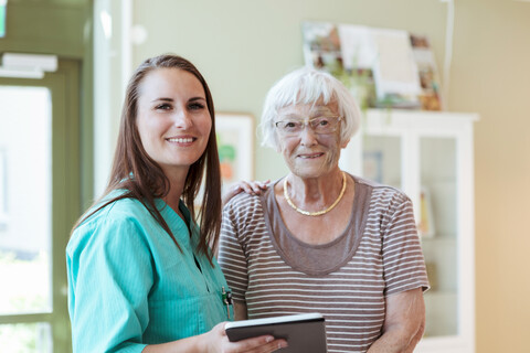 Porträt einer lächelnden älteren Frau und einer Krankenschwester mit digitalem Tablet zu Hause, lizenzfreies Stockfoto