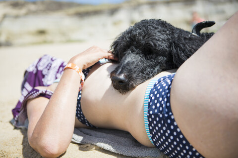 Kopf eines Hundes, der auf dem Bauch einer Frau liegt, die am Strand ein Sonnenbad nimmt, lizenzfreies Stockfoto