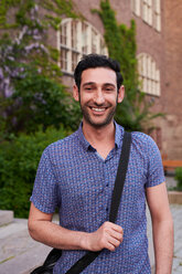 Porträt eines lächelnden jungen Mannes mit Umhängetasche, der vor einer Sprachschule steht - MASF11208