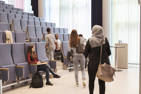 Lächelnde Studenten, die auf ihre Freunde schauen, die auf Stühle im Hörsaal einer High School zugehen, lizenzfreies Stockfoto