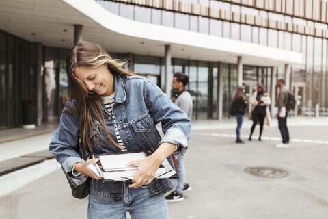Lächelnde junge Frau mit Büchern in der Hand auf dem Universitätscampus, lizenzfreies Stockfoto