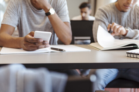 Mittelteil eines männlichen Schülers, der ein Smartphone benutzt, während er am Schreibtisch im Klassenzimmer sitzt, lizenzfreies Stockfoto