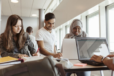 Lächelnde Studenten, die einen Laptop benutzen, während sie mit Freunden am Schreibtisch in der Cafeteria lernen - MASF11335