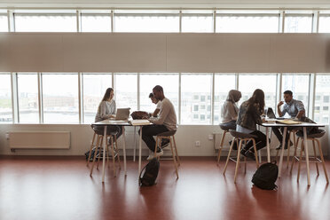 Full length of multi-ethnic students studying at desks by window in high school cafeteria - MASF11338