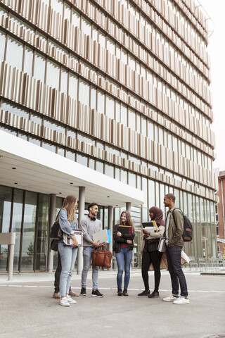 Multiethnische Studenten im Gespräch, während sie gegen ein Gebäude auf dem Universitätscampus stehen, lizenzfreies Stockfoto