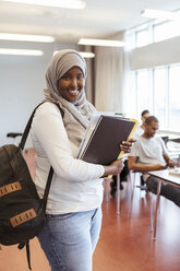 Portrait of smiling woman in hijab standing with books at university classroom - MASF11350