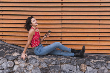 Young woman sitting on a wall listening music with smartphone and headphones - KKAF03114