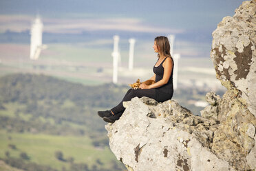 Junge Frau mit Schoßhund sitzt auf einem Felsen und schaut in die Ferne - KBF00503
