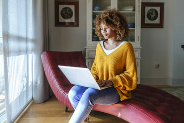 Junge Frau sitzt auf einer Chaiselongue und benutzt einen Laptop zu Hause - KIJF02272
