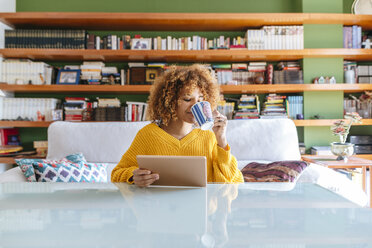 Young woman with curly hair using tablet and drinking coffee at home - KIJF02276
