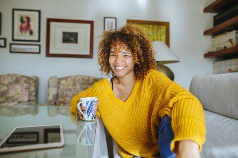 Porträt einer glücklichen jungen Frau mit lockigem Haar, die zu Hause eine Tasse hält, lizenzfreies Stockfoto
