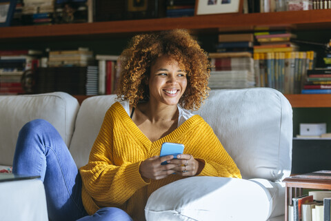 Happy young woman with cell phone sitting on sofa at home stock photo