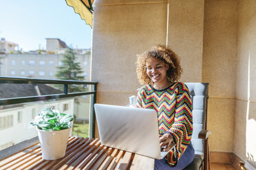 Junge Frau mit lockigem Haar sitzt auf dem Balkon und benutzt einen Laptop - KIJF02291