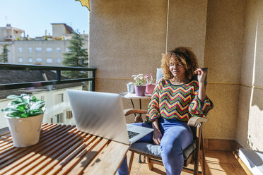 Junge Frau mit lockigem Haar sitzt auf dem Balkon und benutzt einen Laptop - KIJF02292