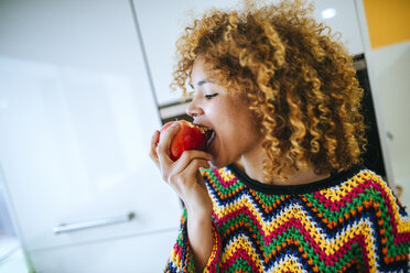 Young woman with curly hair eating an apple in kitchen - KIJF02295
