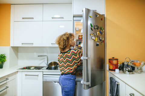 Young woman with curly hair opening the fridge in kitchen stock photo