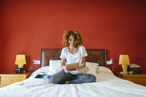 Junge Frau mit lockigem Haar sitzt auf dem Bett und benutzt ein Tablet, lizenzfreies Stockfoto