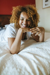 Porträt einer glücklichen jungen Frau mit lockigem Haar, die zu Hause auf dem Bett liegt - KIJF02303