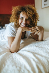 Portrait of happy young woman with curly hair lying on bed at home - KIJF02303