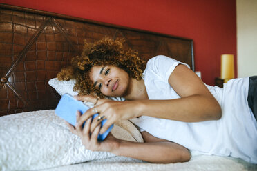 Young woman with curly hair lying in bed at home using cell phone - KIJF02310