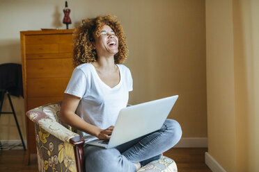 Happy young woman with curly hair sitting in armchair at home using laptop - KIJF02312