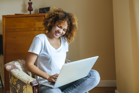 Glückliche junge Frau mit lockigem Haar sitzt zu Hause im Sessel und benutzt einen Laptop, lizenzfreies Stockfoto