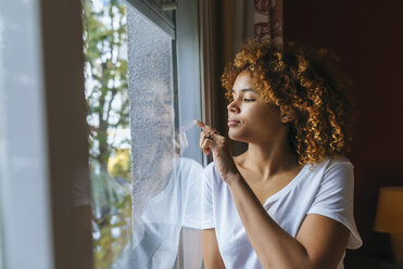 Young woman with curly hair writing with her finger on windowpane - KIJF02316