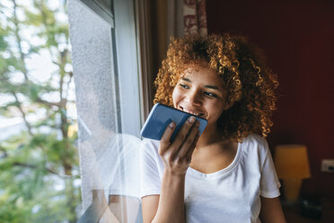 Young woman with curly hair sending voice message with cell phone next to the window - KIJF02317