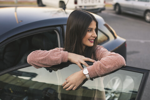 Lächelndes Teenager-Mädchen lehnt sich an die Autotür und schaut in die Ferne, lizenzfreies Stockfoto