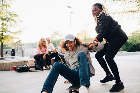 Teenager-Mädchen schiebt glücklichen Mann auf Skateboard beim Spielen im Park, lizenzfreies Stockfoto