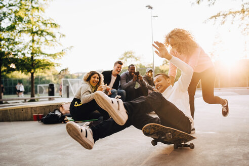 Lachende Freunde fotografieren einen Mann, der vom Skateboard fällt, während eine Frau ihn im Park anschiebt - MASF11372