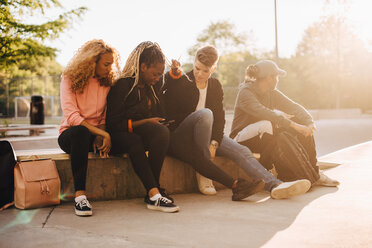 Multi-ethnic friends looking at smart phone while sitting at skateboard park - MASF11379