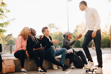 Happy friends greeting man with skateboard at park - MASF11380