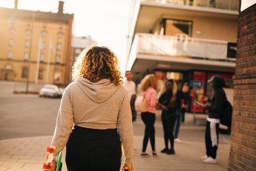 Rear view of woman with skateboard walking towards friends in city - MASF11397