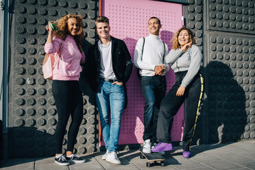 Full length of smiling male and female friends standing against wall on sunny day - MASF11409