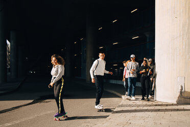 Freunde betrachten Mann und Frau beim Skateboardfahren auf der Straße an einem sonnigen Tag - MASF11413